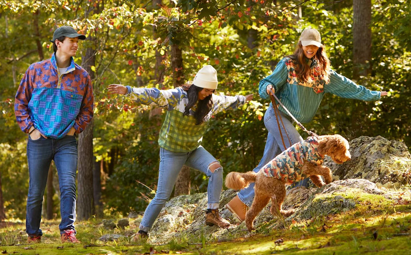 Three people are walking in the woods. The person at left is walking through the grass, the two people on the left are walking over large rocks along with a goldendoodle. The people are wearing jeans and quarter-button fleece pullovers in bright designs.