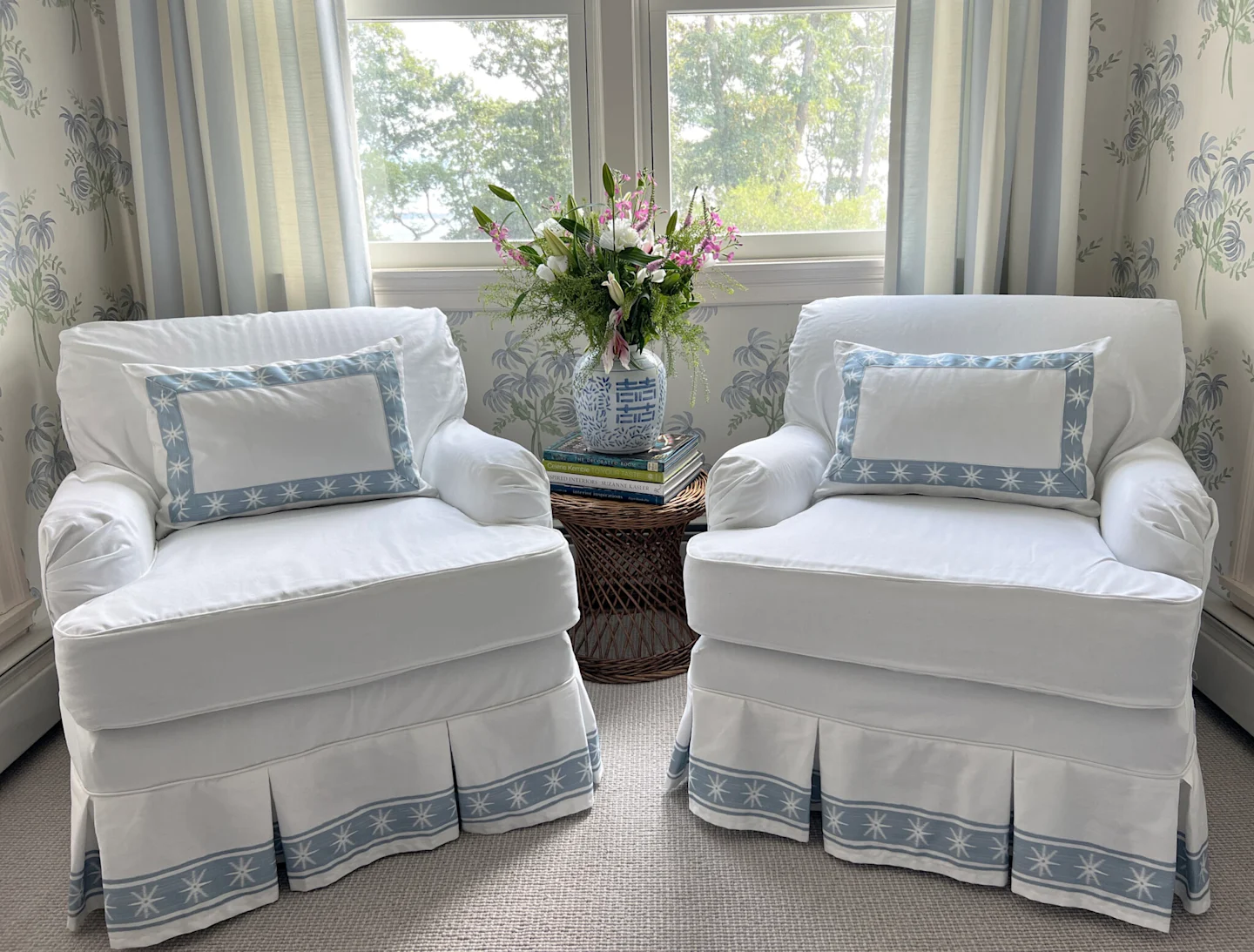 Two white pillows with blue fabric trim around the edges with white stars sit on two chairs covered by white slipcovers with the same fabric trim at the bottom of the chairs’ dust ruffles. The chairs sit in a living room corner with a window behind them t