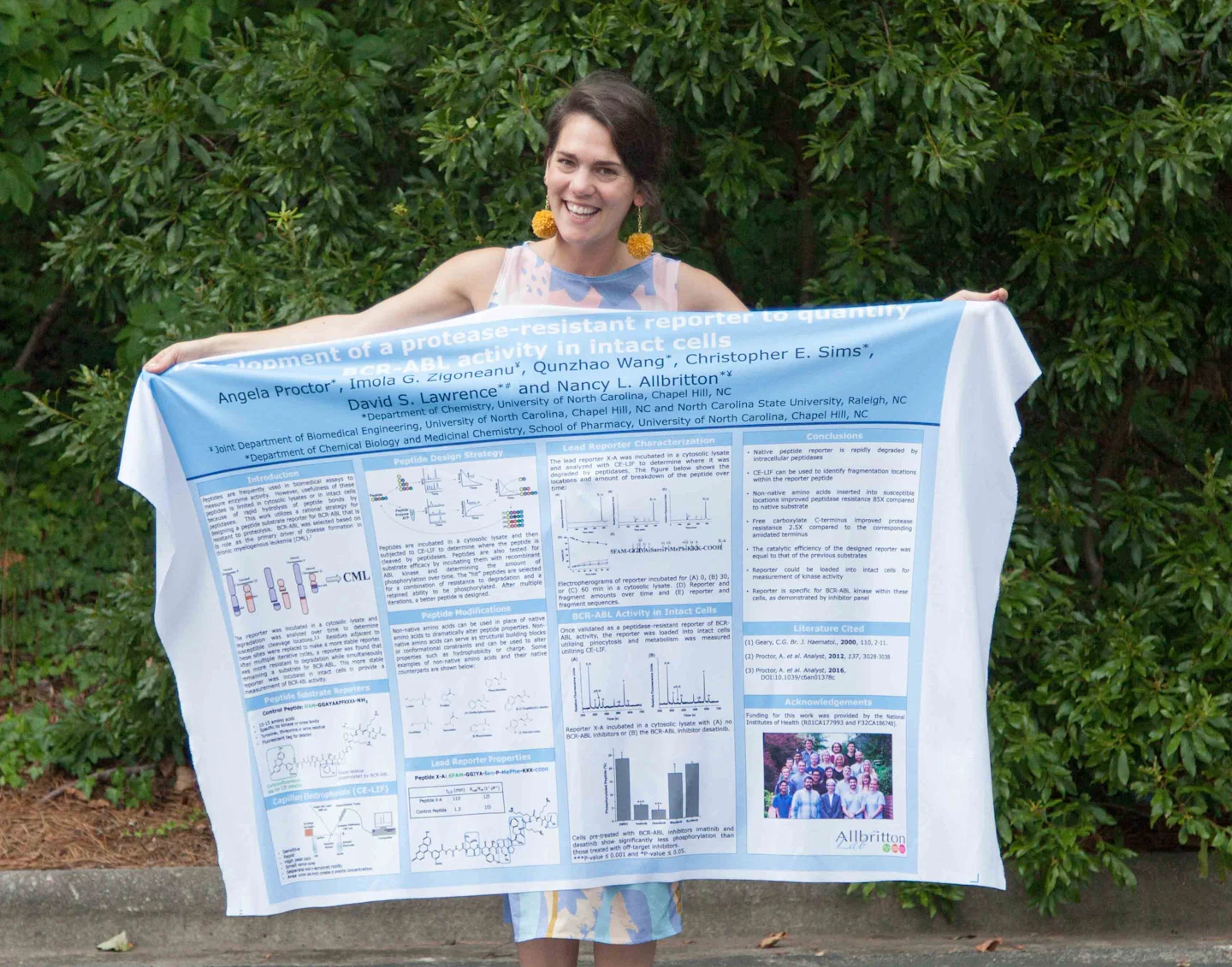 A woman stands in front of green bushes. She is smiling and holding up a fabric poster on which her research has been printed.